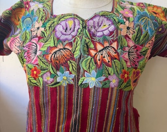 May include: A short-sleeved blouse featuring a deep red base with vertical multicolored stripes. The upper chest and shoulder area are adorned with dense, vibrant floral embroidery in shades of purple, orange, pink, blue, and green.