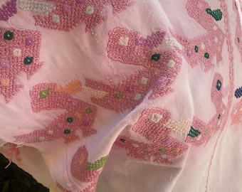 May include: A light pink embroidered blouse with intricate floral patterns in shades of pink, green, and white. The embroidery is detailed and textured, covering the shoulders and sleeves. The blouse has a round neckline and a soft, flowing fabric.