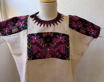 May include: A cream-colored huipil featuring intricate hand-embroidered geometric patterns in black, purple, orange, and blue. The garment has a round neckline with dark brown decorative stitching and wide sleeves with matching embroidery.