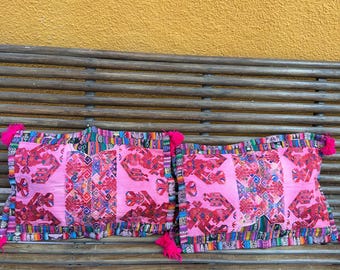 May include: Two pink decorative pillows with intricate red and multicolored patterns. Each pillow has a border of colorful fabric and pink tassels on the corners. The pillows are placed on a wooden bench against a yellow wall.