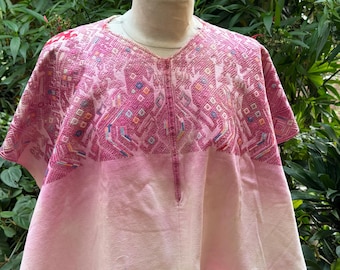 May include: A pink, short-sleeved tunic with a V-neck. The top portion features a detailed geometric pattern in shades of pink, red, and blue. The lower part of the tunic fades to a lighter pink. The garment is displayed on a mannequin.