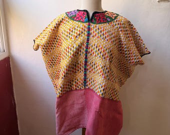 May include: A handmade, colorful tunic displayed on a wooden mannequin. The top portion features a yellow, patterned fabric with a diamond design, accented with a floral embroidered collar. The bottom is a solid red, vertically striped fabric.