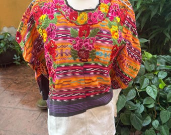 May include: A short-sleeved blouse featuring a vibrant, multi-colored woven pattern with horizontal stripes in orange, pink, and purple. The neckline and chest are adorned with intricate, dense floral embroidery in shades of red, yellow, and pink.