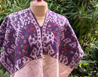 May include: A purple and white embroidered poncho with a V-neck. The poncho features a detailed pattern with geometric shapes and animal figures in various colors. The lower section is a solid light pink color.