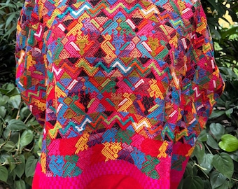 May include: A vibrant red blouse featuring intricate, multi-colored geometric embroidery and stylized bird motifs. The garment has a round neckline with a black border and a wide, solid red hem at the bottom, showcasing a dense, woven textile pattern.