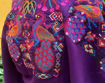 May include: A vibrant purple embroidered blouse featuring colorful bird and geometric designs. The intricate embroidery includes shades of orange, blue, pink, and green. The blouse has long sleeves and a round neckline, showcasing traditional craftsmanship.