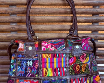 May include: A colorful patchwork handbag with brown leather accents. The bag features a variety of vibrant woven fabric squares in shades of red, blue, yellow, and pink. It has two brown leather handles and a zipper closure.