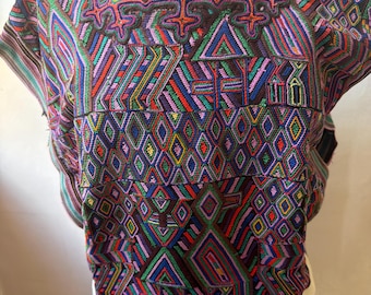 May include: A colorful, embroidered top with a square neckline. The fabric features geometric patterns in shades of purple, blue, green, red, and yellow. The bottom portion is white with a black trim.