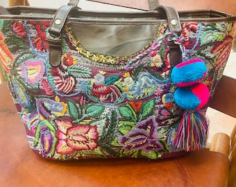 Pode incluir: Uma bolsa tote bordada colorida com alças de couro marrom e um tassel com pompons azuis e rosa.
