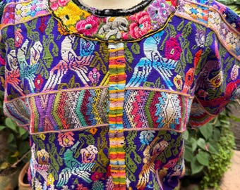 May include: A vibrant purple blouse featuring intricate, multi-colored embroidery throughout. The design includes detailed floral patterns, stylized bird motifs, and geometric shapes. A central vertical panel displays a rainbow-striped texture.