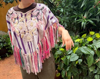 May include: A white poncho featuring vertical purple stripes, intricate floral embroidery in shades of pink and cream, and decorative butterfly motifs. The garment is finished with long, multi-colored fringe trim along the bottom hemline.