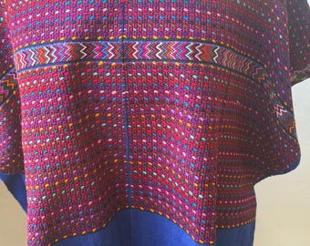 May include: A woven poncho featuring a deep red upper section with intricate multi-colored geometric patterns and a solid blue lower section. The garment has a rounded neckline with detailed embroidery and short, wide sleeves with patterned edges.