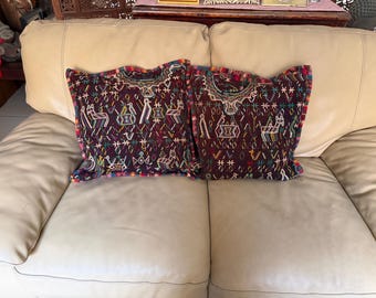 May include: Two decorative throw pillows with intricate embroidered designs. The pillows feature a deep burgundy background with colorful geometric and animal motifs. The edges are trimmed with a vibrant, multi-colored border, adding a pop of color to the design.