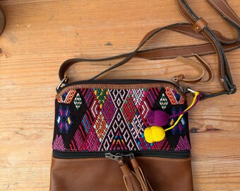 Pode incluir: Bolsa transversal de couro marrom com parte superior em tecido colorido. O tecido apresenta um padrão geométrico em várias cores, incluindo rosa, roxo, amarelo e preto. A bolsa tem uma longa alça marrom e um puxador de zíper com borla.