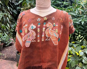 May include: A rust-colored textile garment featuring intricate embroidery of two large birds facing each other, surrounded by smaller geometric patterns. The piece has short sleeves, a V-neckline, and a bottom hem finished with long brown fringe.