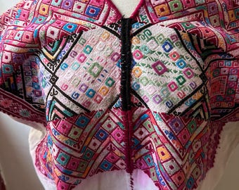 May include: A white top with a vibrant, embroidered yoke. The yoke features a geometric pattern in shades of pink, purple, green, and blue. A black trim runs down the center of the top. The garment is displayed on a mannequin.