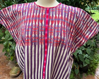 May include: A woven huipil blouse featuring a vibrant purple and white striped pattern on the lower section and a complex geometric design in purple and red on the upper chest. A bright pink vertical trim runs down the center of the garment.