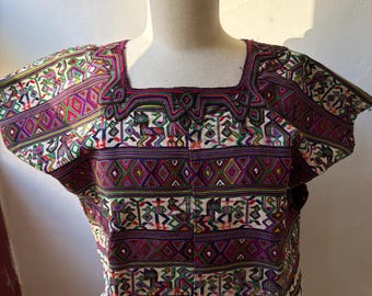 May include: A colorful, square-neck top with short sleeves. The garment features intricate embroidery in shades of purple, green, red, and white, with geometric and figurative patterns. The top is displayed on a mannequin.