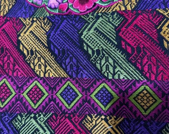 May include: A woven textile garment featuring a vibrant, multi-colored geometric pattern in shades of purple, pink, green, and yellow. The neckline is adorned with a dark red velvet trim and a detailed, colorful floral embroidery design.