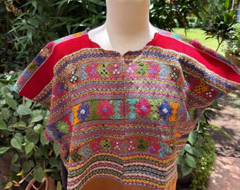 May include: A short-sleeved blouse featuring a vibrant, multi-colored woven pattern with horizontal stripes in shades of red, blue, green, and yellow. The garment has a solid brown hem at the bottom and a central vertical seam down the front.