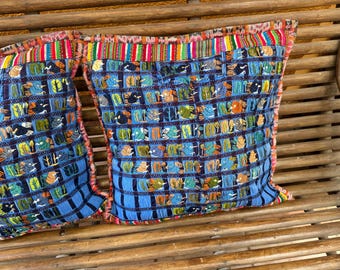 May include: Two blue decorative pillows with a woven design featuring colorful birds and trees. The pillows have a striped border in red, pink, and green. The pillows are resting on a wooden bench.