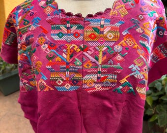 May include: A magenta short-sleeved blouse featuring a scalloped neckline and intricate, multi-colored geometric and floral embroidery across the chest and shoulders. The garment is displayed on a mannequin, showcasing the vibrant thread work.