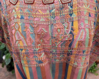 May include: A woven tunic featuring a square neckline with raised, dark red embroidered trim. The upper section displays intricate, multi-colored geometric and stylized animal patterns, while the lower portion consists of vertical stripes in muted tones.