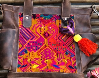May include: A brown leather tote bag with a colorful embroidered panel featuring geometric patterns in shades of pink, orange, yellow, and blue. The bag has a top handle and a detachable shoulder strap.