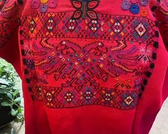 May include: A vibrant red, embroidered tunic with intricate patterns. The neckline and sleeves are adorned with colorful floral and geometric designs. The embroidery features a variety of colors, including blue, green, yellow, and purple, creating a striking visual effect.