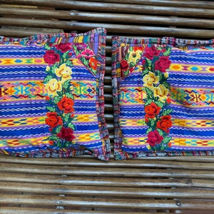 Puede incluir: Dos almohadas cuadradas con un fondo a rayas azul, amarillo y morado. Las almohadas tienen diseños florales bordados con detalles en rojo, amarillo y verde.