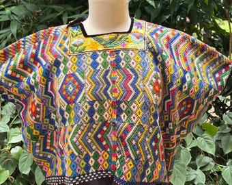 May include: A colorful, hand-embroidered huipil featuring intricate geometric patterns in shades of blue, yellow, green, pink, and red. The square neckline is accented with a yellow panel displaying floral and bird motifs, finished with a black hem.