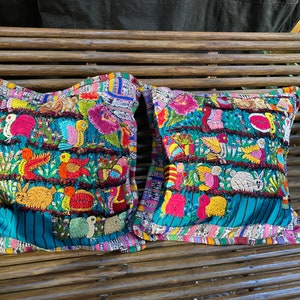 Puede incluir: Dos fundas de cojín bordadas de colores con fondo turquesa y diseños intrincados que representan pájaros, flores y animales. Los cojines se muestran en un banco de madera.