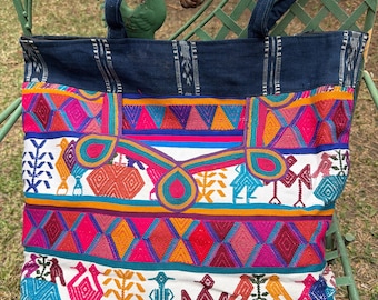 May include: A colorful tote bag with a denim top and straps. The body of the bag features vibrant embroidered patterns in pink, orange, blue, and white. The design includes geometric shapes and bird motifs, creating a unique and eye-catching accessory.