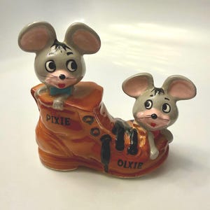 May include: A ceramic figurine featuring two grey mice with pink ears and black whiskers emerging from a brown shoe. The shoe has black laces and the word PIXIE printed on the side, while the word OLXIE is printed near the heel area.