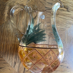 May include: A clear glass pitcher with a pineapple design. The pitcher has a handle and is decorated with a yellow and orange pineapple pattern with green leaves.