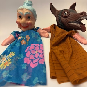 May include: Two vintage hand puppets. One is a smiling elderly woman with blue hair and a blue floral dress. The other is a brown wolf or dog with an open mouth and a brown striped shirt.