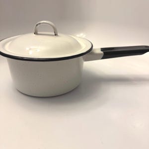 May include: A white enamel saucepan with a speckled finish, featuring a matching lid with a small handle and a long, straight black handle. The rim of the pan and the lid are accented with a thin black border, creating a clean, functional design.