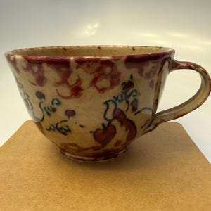 May include: A handmade ceramic teacup with a wide rim and a handle. The cup features a beige base with red and brown floral patterns and a red rim. The cup is sitting on a brown surface.