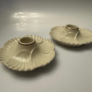 May include: Two off-white ceramic candle holders. Each holder has a central cup with a gold rim, set on a scalloped, petal-like base. The holders are designed to hold candles, adding a decorative touch to any space.
