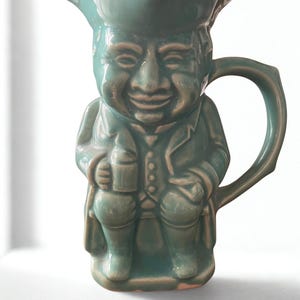 May include: A teal ceramic pitcher shaped like a smiling man in a tricorn hat and period clothing. The handle is curved and the pitcher is glazed with a glossy finish. The man is holding a small mug.