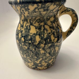 May include: A yellow ceramic pitcher with a blue and white marbled design. The pitcher has a wide mouth and a handle.