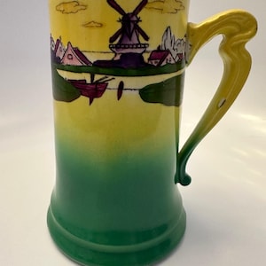 May include: A green and yellow ceramic mug with a scene of a windmill and houses painted on the side. The handle is yellow and has a unique shape.