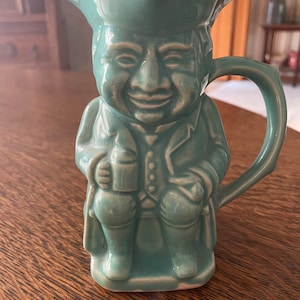 May include: A ceramic pitcher shaped like a man wearing a hat and holding a mug. The pitcher is a light teal color.