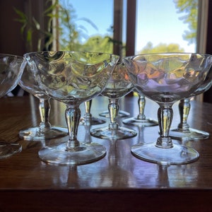 May include: A set of clear glass champagne coupes with a textured design. The glasses are arranged in a row on a wooden table.