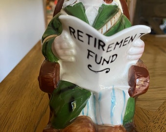 Pode incluir: Uma figurinha de cerâmica de um homem sentado em uma cadeira de balanço, lendo um jornal que diz "Fundo de pensões". O homem está vestindo uma jaqueta verde, calças listradas azul e branco e tem um cachimbo na boca.