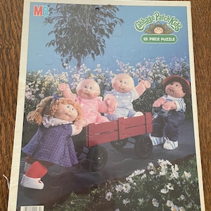 May include: A 25-piece jigsaw puzzle featuring four Cabbage Patch Kids dolls. The dolls are dressed in colorful outfits and are riding in a red wagon. The puzzle is designed for ages 3 to 7.