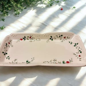 May include: A rectangular, light pink serving platter with a scalloped edge. The platter is decorated with a delicate floral pattern of green leaves and red berries. The design is repeated around the edges of the platter.