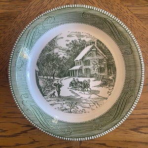 May include: A round, white and green decorative plate with a winter scene illustration. The plate features a green border with a repeating scroll design and a white rim with small, raised dots. The central image depicts a house, trees, and figures.