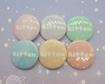 Könnte beinhalten: Sechs pastellfarbene runde Knöpfe mit dem Wort "Kitten" in weißen Buchstaben auf jedem. Die Knöpfe haben ein dezentes Muster aus weißen Pfotenabdrücken.