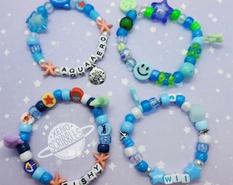 May include: Four colorful beaded bracelets with different charms and letter beads spelling out the words "AQUA AERO", "FISHY", "WILL", and a smiley face. The bracelets are on a light blue background.